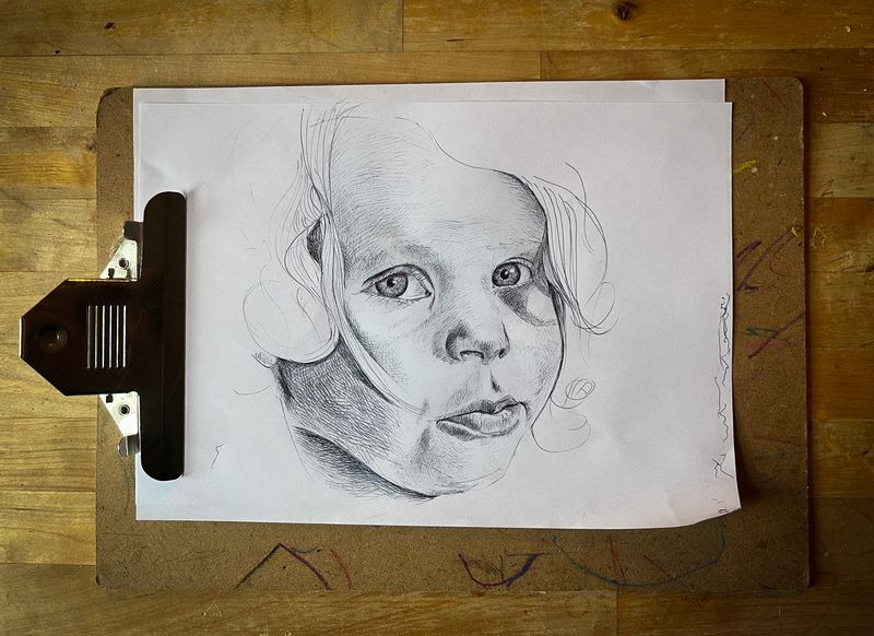 Black bic biro crosshatch drawing of a little boy’s face looking a bit cheeky. Set on a wooden A4 clipboard lying on a wooden table