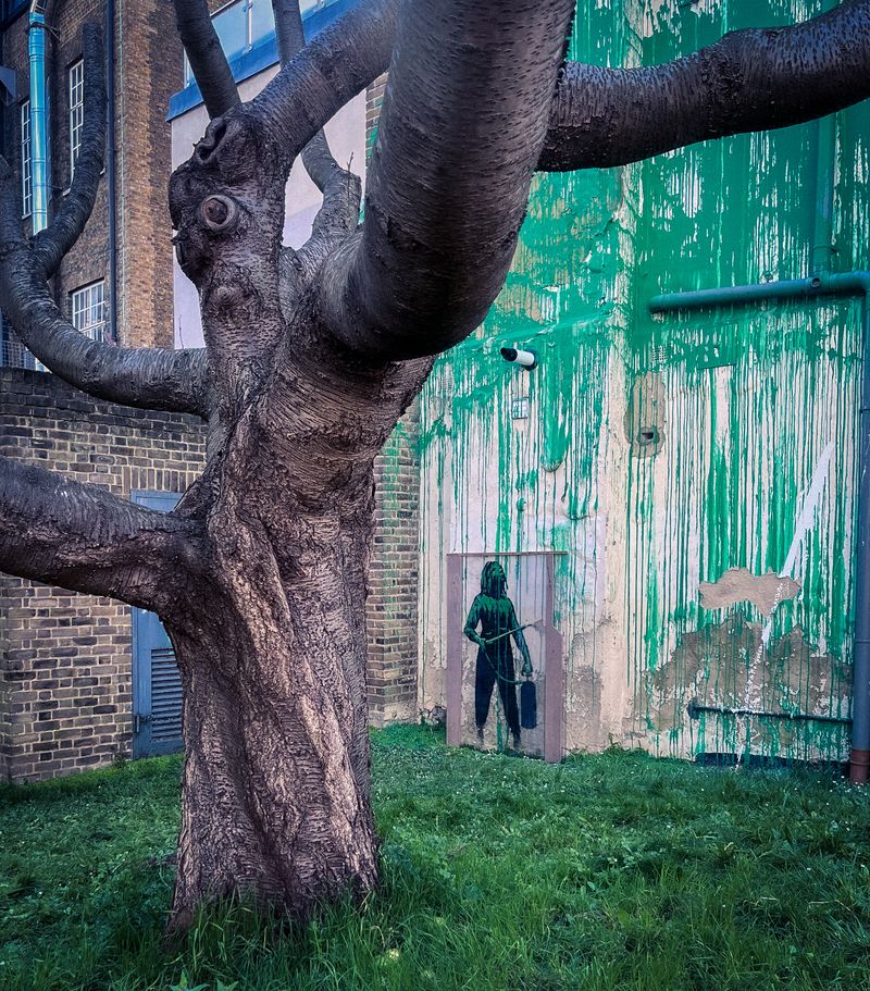 stencil of a person in green spraying green foliage with a pressure hose onto a wall at the end of a terraced block with a dead tree in the foreground